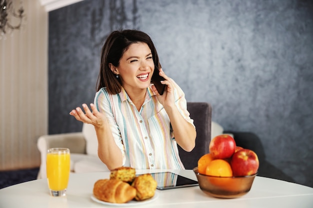 Mulher feliz sentado à mesa de jantar de manhã e usando o telefone. mulher ocupada, tendo uma ligação com o chefe.