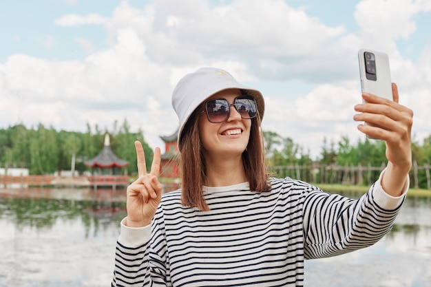 Mulher feliz sorridente usando camisa listrada panamá óculos de sol em pé perto do lago fazendo selfie mostrando sinal v tendo videochamada viajando desfrutando de passeios turísticos