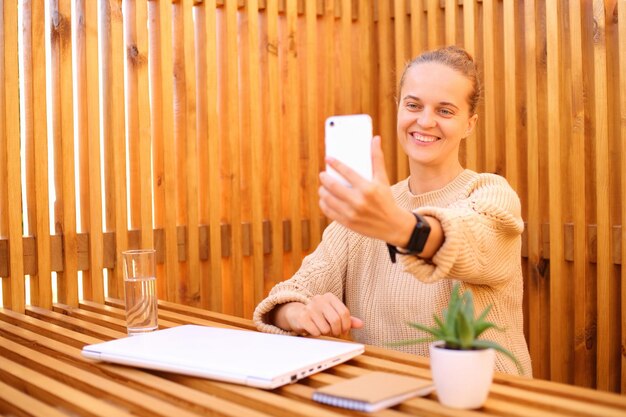 Mulher feliz sorridente, vestindo camisa branca, sentada no café ao ar livre, trabalhando no laptop, tendo uma pausa para uma chamada de vídeo no smartphone, expressando emoções positivas