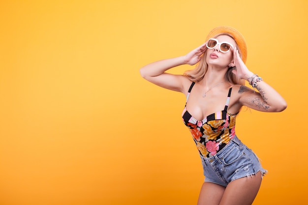 Mulher feliz sorrindo com chapéu e óculos posando sobre fundo amarelo em estúdio