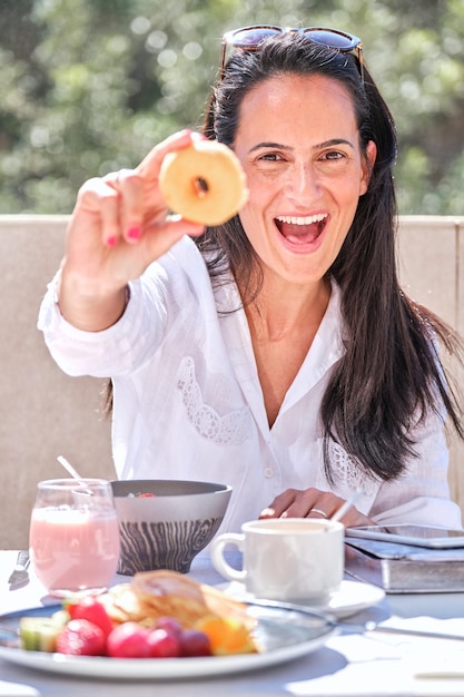 Mulher feliz tomando café da manhã com iogurte e frutas e bagas mostrando donuts saborosos e olhando para a câmera enquanto está sentada na mesa na varanda do hotel em um dia ensolarado