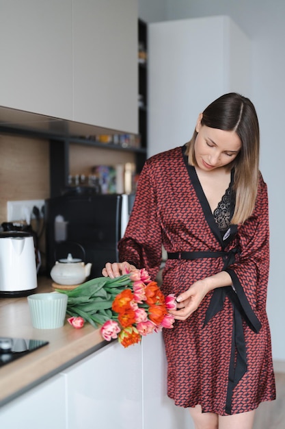 Mulher feliz tomando um café em casa na cozinha vestindo um roupão de seda enquanto desfruta de flores frescas