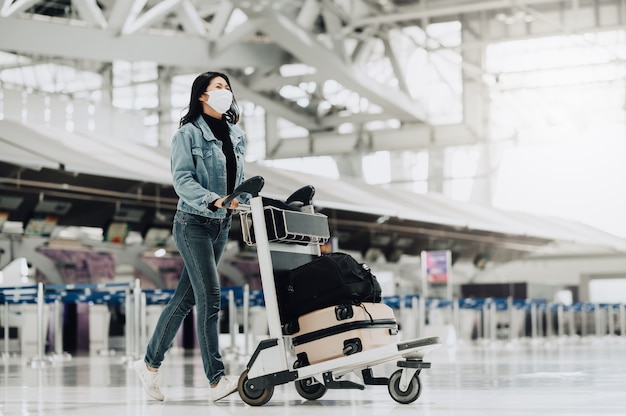 Mulher feliz turista asiática com máscara andando com carrinho de bagagem