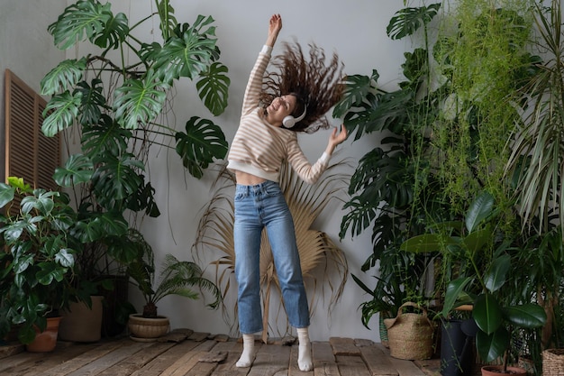 Foto mulher feliz usa fones de ouvido sem fio ouvindo música dançando no jardim de casa com plantas tropicais