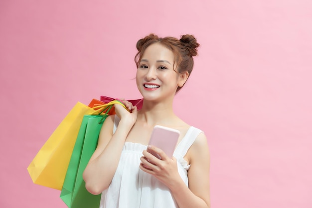 Mulher feliz usando smartphone e segurando sacolas de compras isoladas em rosa