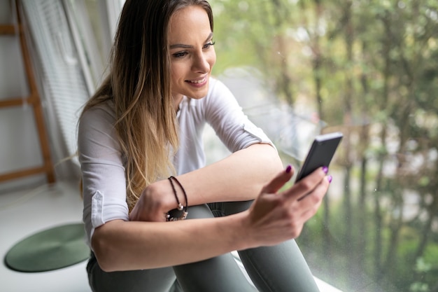 Mulher feliz usando telefone celular relaxando pela janela em casa. conceito de vício online de saúde mental.