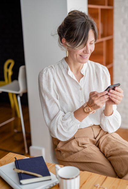 Mulher feliz usando um telefone celular enquanto trabalha em casa. Bela dama navegando no site do telefone