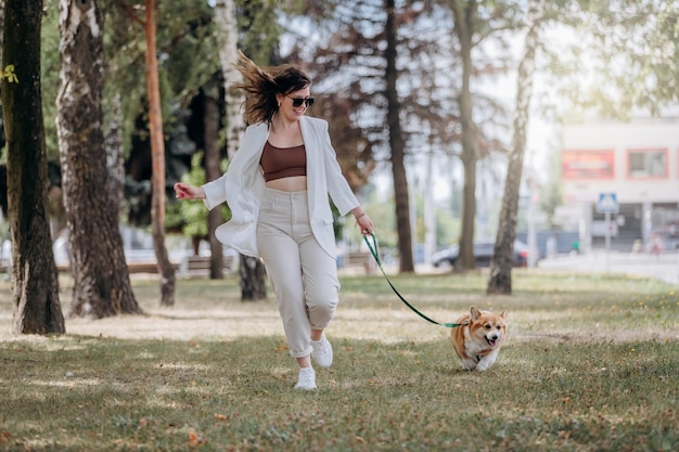 Mulher feliz vestindo terno branco e óculos de sol andando no parque da cidade com cachorro Welsh Corgi Pembroke