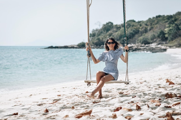 Mulher feliz viajante em vestido xadrez branco e preto e óculos de sol pretos relaxantes no balanço. Praia turística do mar Tailândia, Ásia, viagem de viagem de férias de férias de verão. Phuket. Tailândia.