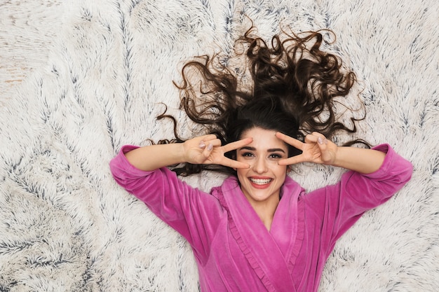 Mulher feminina com cabelo longo encaracolado, vestindo um roupão feminino deitada sobre pelo branco no apartamento