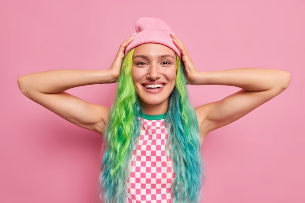 Foto mulher fica com as mãos na cabeça feliz por comprar roupa nova tem tintas para o cabelo colorido tem piercing no nariz isolado no rosa