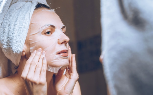 Mulher fica perto de um espelho com uma toalha na cabeça e coloca uma máscara de cosmética