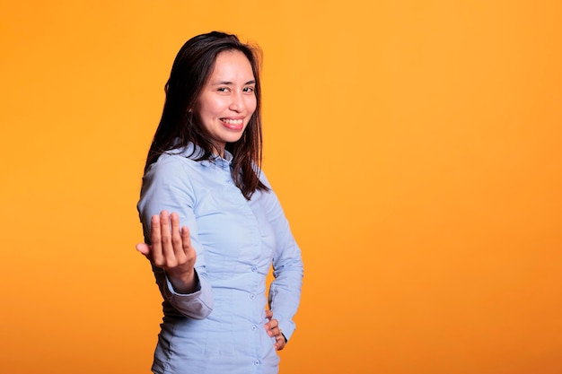 Mulher filipina alegre positiva fazendo gesto, acenando com o dedo posando no estúdio sobre fundo amarelo. Modelo sorridente convidando você para uma conversa confidencial. símbolo de chamada