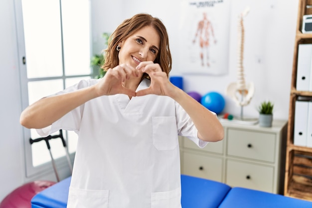 Mulher fisioterapeuta de meia-idade trabalhando na clínica de recuperação de dor sorrindo apaixonadamente fazendo forma de símbolo de coração com as mãos. conceito romântico.