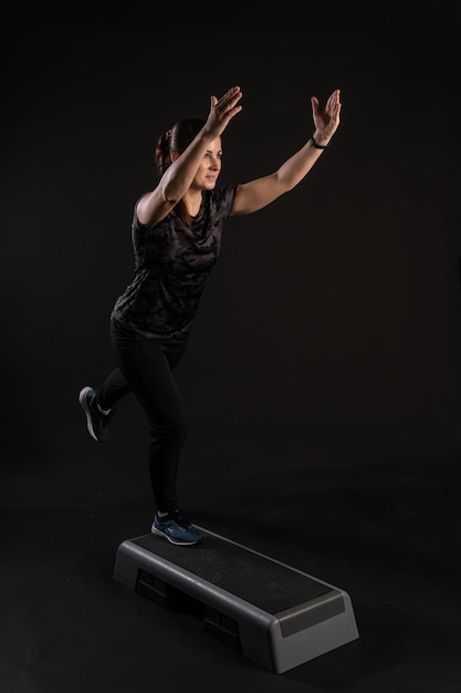 Mulher fitness em uma plataforma de degraus fica em um rack Entra para esportes em um fundo preto olha para