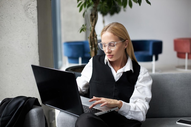 Mulher focada e confiante em óculos usando um laptop para trabalho remoto sentado no sofá