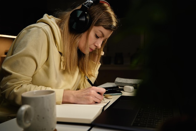 Mulher focada em roupas casuais usando fones de ouvido olhando para a tela do laptop e desenhando ilustração