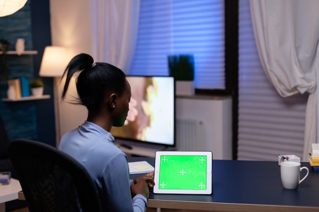 Mulher freelance africana, desfrutando de trabalhar em casa, olhando para o tablet pc com espaço de cópia disponível na mesa. Usando computador de exibição de chave de croma de maquete.