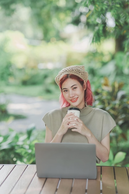 Mulher freelance asiática segurando uma xícara de café quente sorrindo digitando no teclado e trabalhando laptop na mesa de madeira no café Mulher empresária trabalhando para seus negócios no café Trabalho de negócios em qualquer lugar conceito