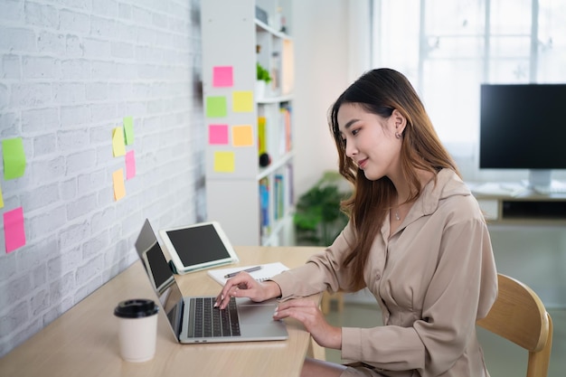 Mulher freelance asiática sorrindo digitando no teclado e trabalhando no laptop na mesa de madeira em casa Mulher empresária trabalhando para seus negócios na sala de estar em casa Conceito de trabalho de negócios em casa