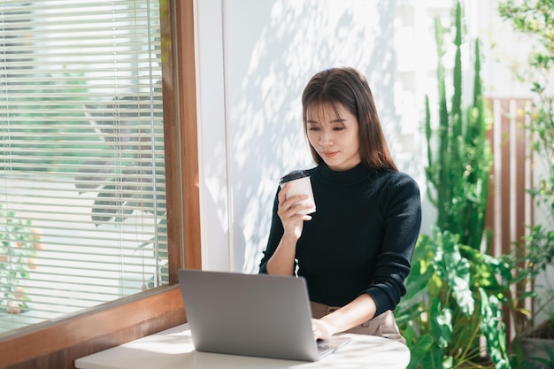 Mulher freelance asiática sorrindo segurando uma xícara de café quente e trabalhando no computador portátil na mesa de madeira no café Mulher empresária trabalhando para seus negócios na cafeteria Trabalho comercial de qualquer lugar