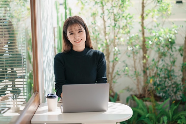 Mulher freelance asiática sorrindo toma uma xícara de café quente e trabalhando no laptop na mesa de madeira no café Mulher empreendedora trabalhando para seu negócio na cafeteria Trabalho de negócios de qualquer lugar