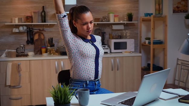 Mulher freelance esticando os braços por causa da exaustão enquanto trabalhava horas extras em casa. Funcionário concentrado e ocupado usando rede de tecnologia moderna sem fio à noite lendo, escrevendo, pesquisando