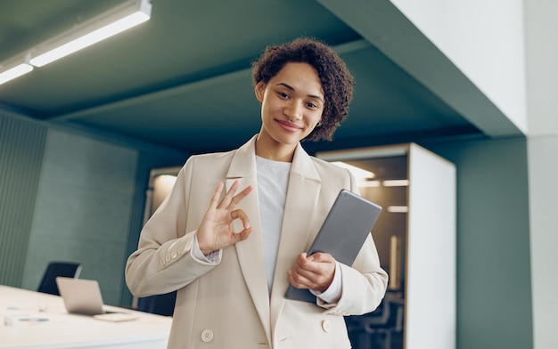 Mulher freelancer com tablet digital de pé em um fundo de coworking moderno e mostrando o sinal ok