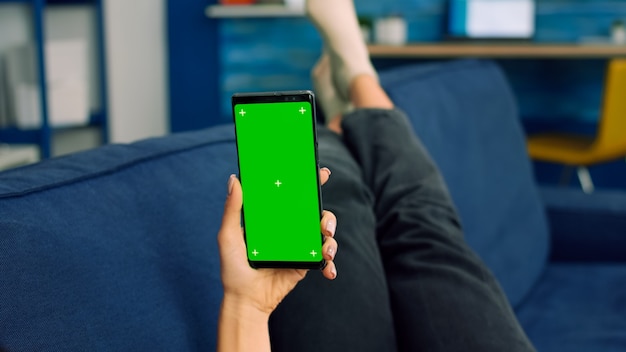 Mulher freelancer deitada no sofá, olhando para a tela verde do chroma key do smartphone no modo vertical