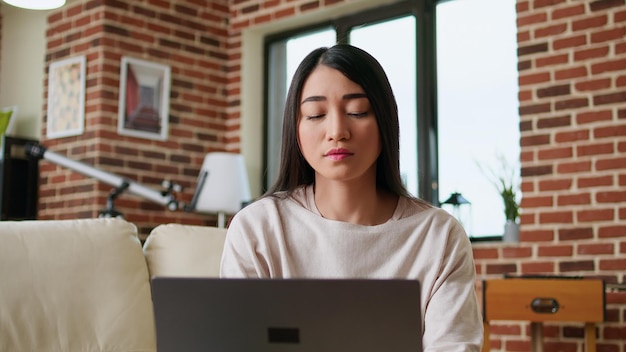 Mulher freelancer focada trabalhando remotamente em casa no laptop moderno. Linda jovem adulta fazendo trabalho de freelancer remoto no computador portátil enquanto está sentado no sofá dentro do apartamento.