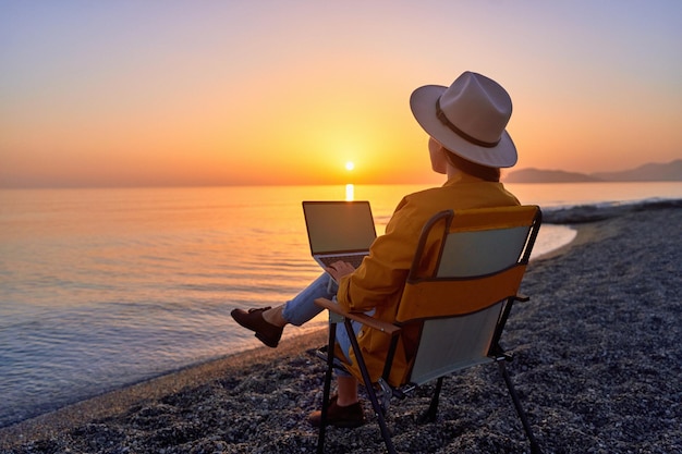 Mulher freelancer milenar livre satisfeita usando computador e sentado na praia pelo mar calmo ao pôr do sol apreciação do conceito de trabalho remoto do escritório dos sonhos