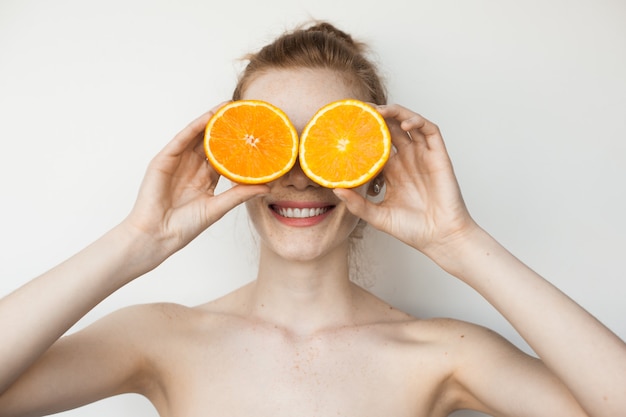 Mulher gengibre com sardas cobrindo os olhos com fatias de laranja e sorrindo em uma parede branca com ombros nus