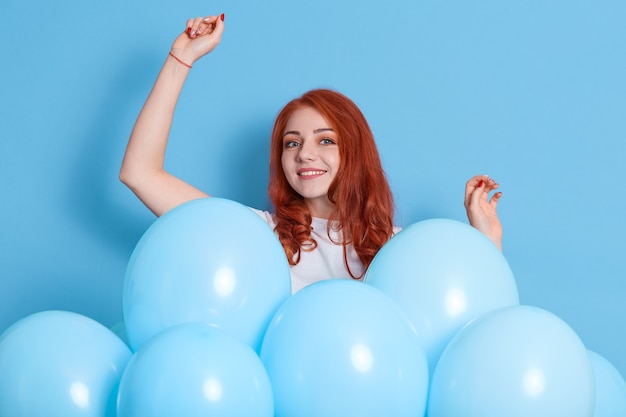 Mulher gengibre de expressão alegre, levanta a mão, dança ao som de música, se diverte na festa de aniversário, estando rodeada de balões azuis, tem bom humor no aniversário, menina comemora feriado.