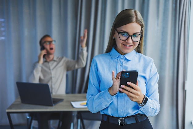 Mulher gerente de óculos clássicos sorrindo durante o horário de trabalho no escritório falando na mesa do telefone com colegas de laptop em segundo plano Um colega está no foco seletivo de fundo