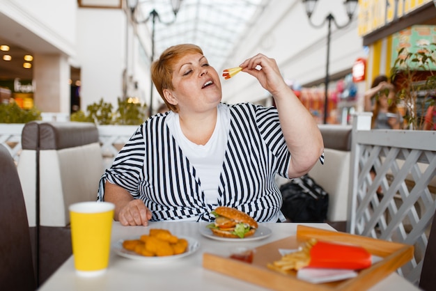 Mulher gorda comendo hambúrguer na praça de alimentação do shopping.