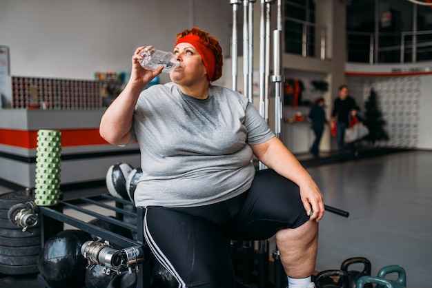 Mulher gorda e suada bebe água após um treino ativo no ginásio. Queima de calorias, mulher obesa fazendo exercícios no clube