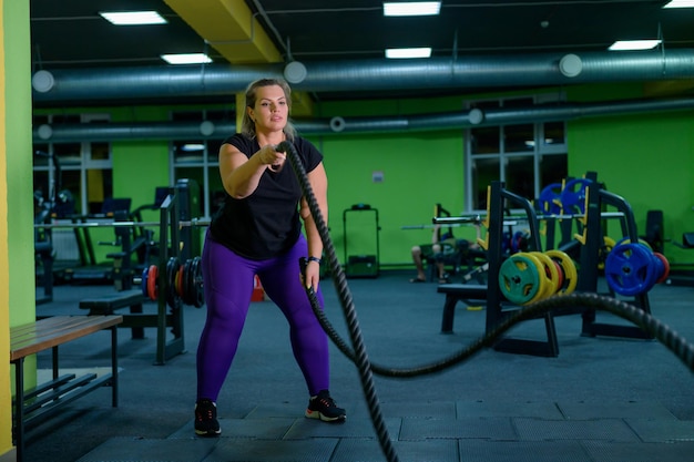 Mulher gorda fazendo treinamento de força usando cordas de batalha no ginásio A menina obesa está envolvida em exercícios circulares para perda de peso