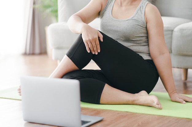 Mulher gorda gordinha asiática madura sentada no chão na sala de estar prática aula de ioga online com o computador. mulher tendo aula de treinamento de meditação no laptop.