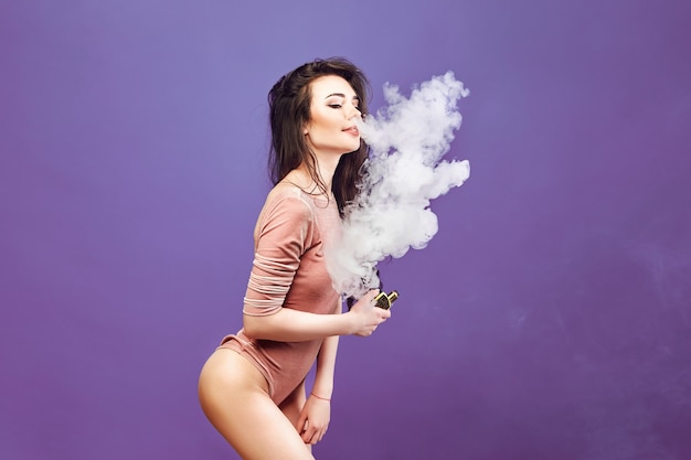 Mulher gostosa em body em pé e vaping na parede roxa.