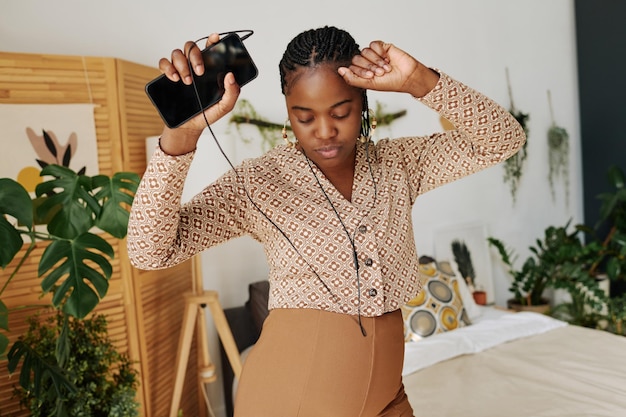 Mulher grávida africana em fones de ouvido dançando no quarto enquanto ouve música em seu celular