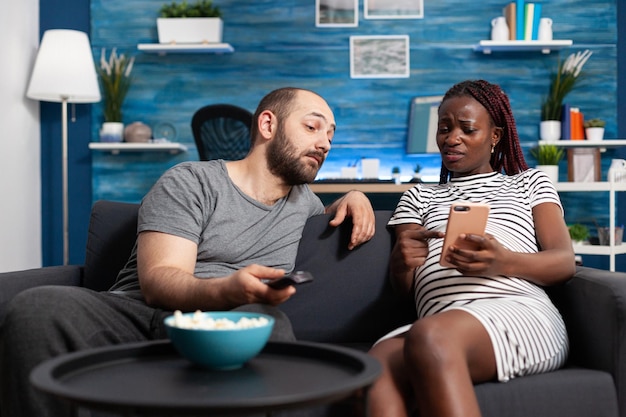 Mulher grávida afro-americana preocupada sentada no sofá em casa enquanto mostrava ao homem caucasiano curioso algo no smartphone. Casal jovem multiétnico olhando contas emitidas erradas usando telefone moderno.