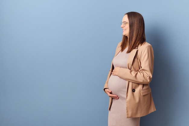 Mulher grávida alegre, vestida com vestido e casaco, isolada sobre um fundo azul, acariciando a barriga, olhando de lado para o espaço de cópia para publicidade ou texto promocional