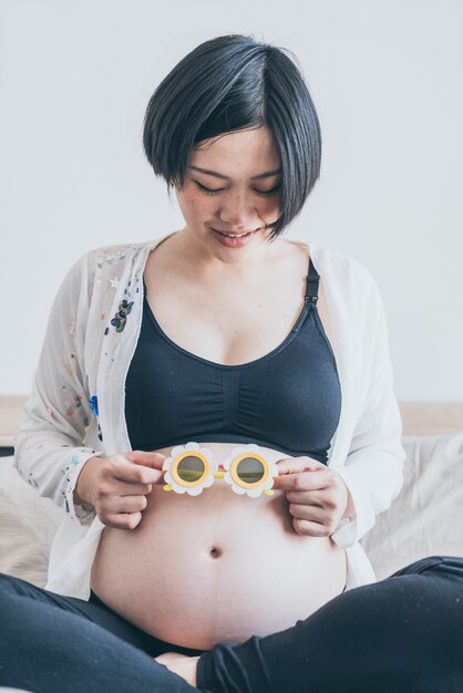 Mulher gravida asiática bonita nova com os óculos de sol da flor em sua barriga em casa