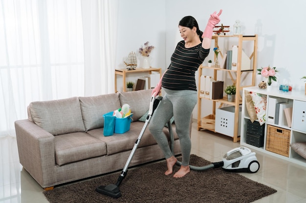 mulher grávida asiática enérgica de corpo inteiro levantando a mão está se divertindo ouvindo músicas com movimentos de dança enquanto aspira o chão da sala de estar em casa.