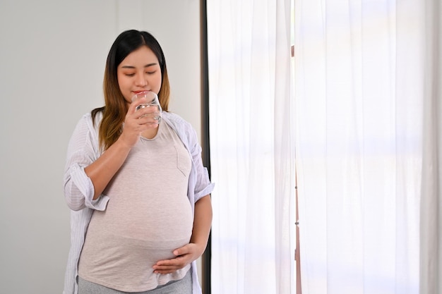 Mulher grávida asiática feliz fica perto da janela bebendo água e tocando sua barriga