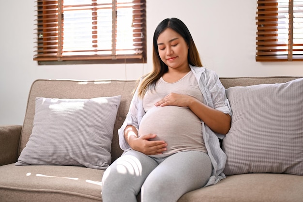 Mulher grávida asiática feliz relaxa no sofá em sua sala de estar tocando a barriga com amor