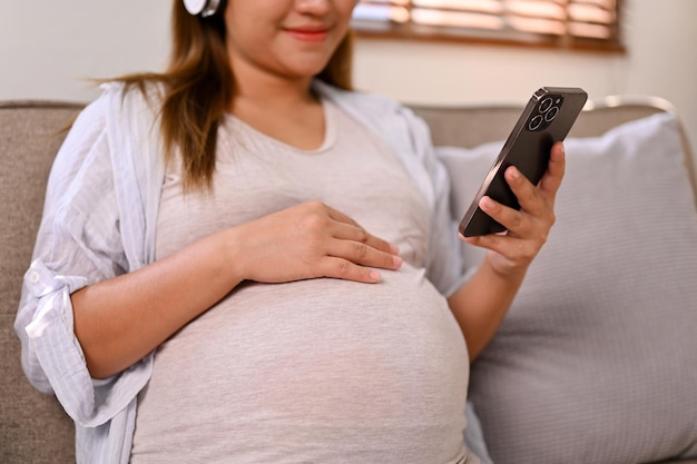 Mulher grávida asiática feliz usando fones de ouvido usando seu smartphone relaxando no sofá