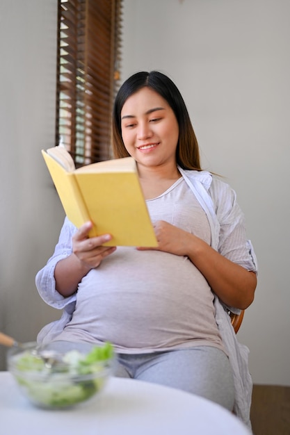 Mulher grávida asiática relaxada lendo um livro em uma mesa de jantar se preparando para ser mãe