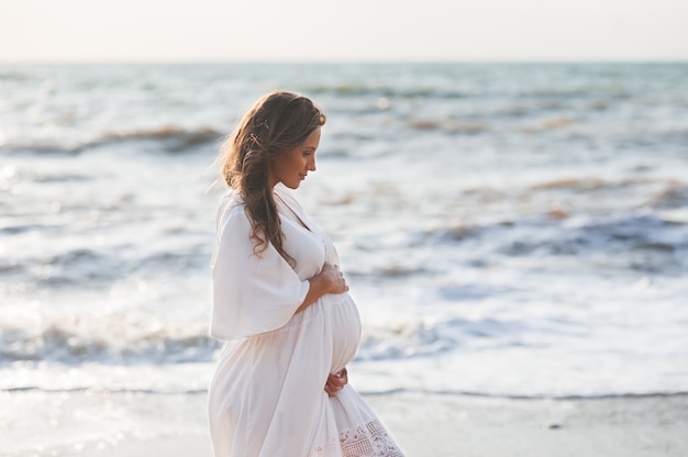 Mulher grávida bonita e elegante com um vestido branco moderno sobre o mar