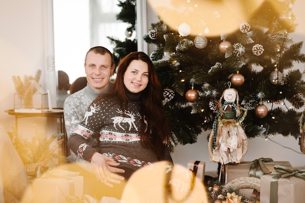 Mulher grávida bonita e homem em suéteres macios perto da árvore em casa Feliz Ano Novo